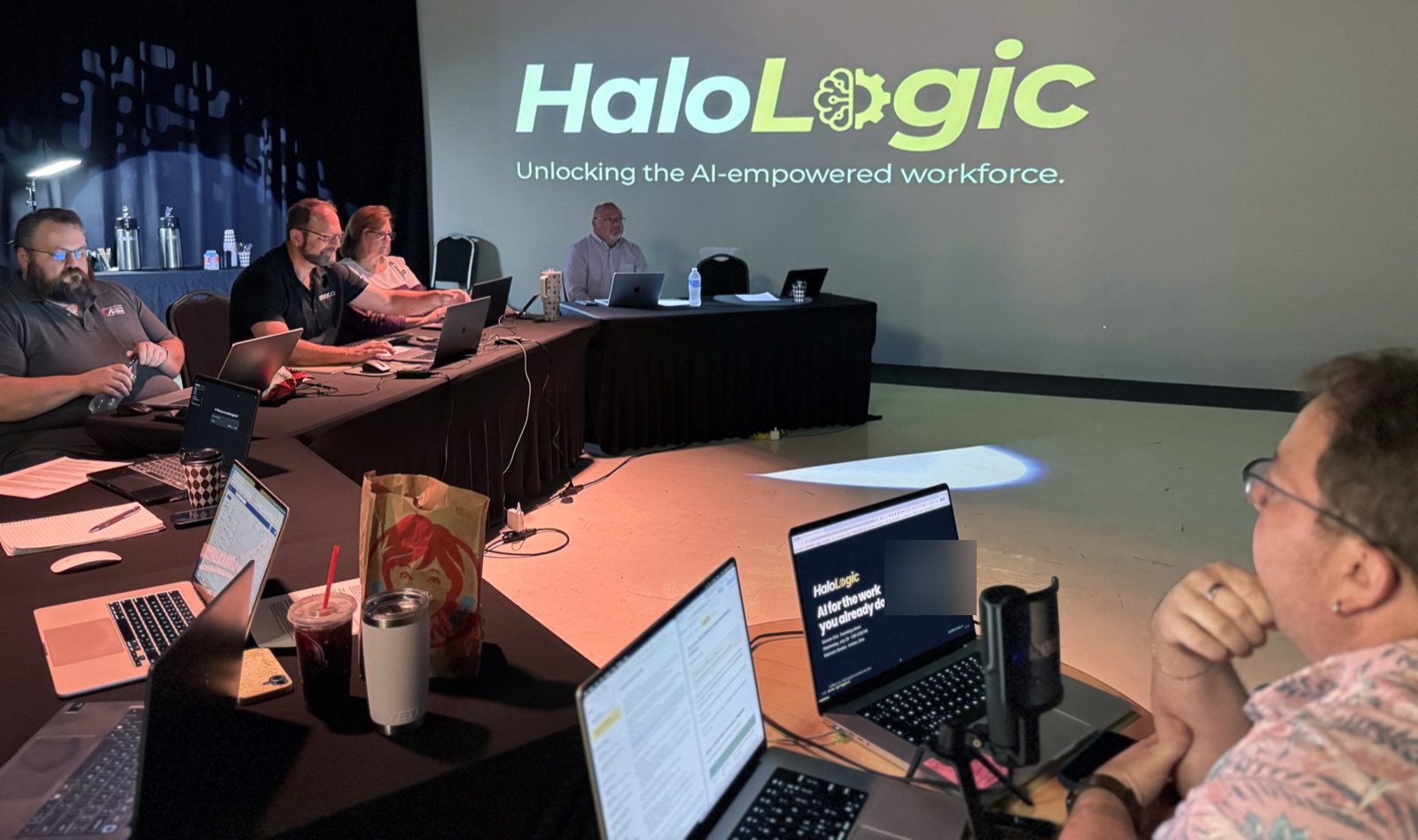 Wide view of the workshop room: attendees at laptops around U-shaped tables, Halo Logic projected on the wall.